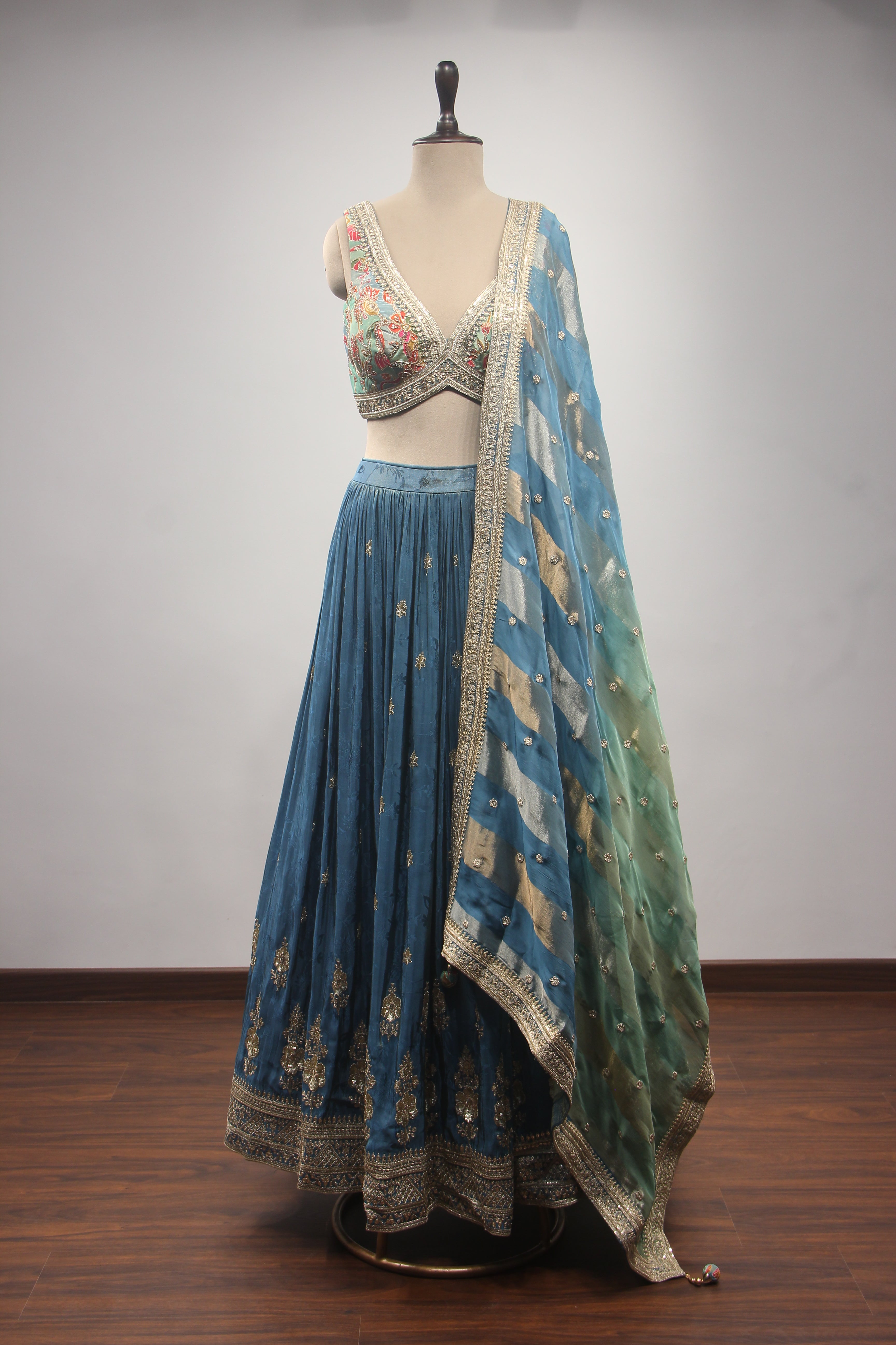 A blue and green embroidered traditional Indian lehenga choli set displayed on a mannequin. The set features a blue pleated skirt with gold embroidery, a matching embroidered blouse, and a sheer green dupatta with gold motifs.