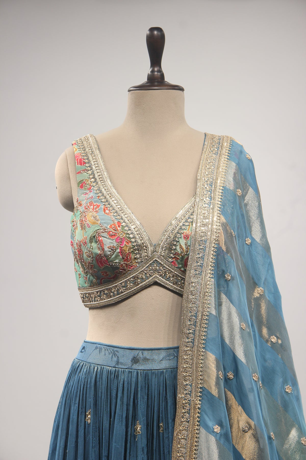 A traditional Indian ethnic wear set displayed on a mannequin, featuring a light blue pleated skirt, a heavily embroidered light blue blouse with gold borders, and a matching sheer blue dupatta with gold motifs.